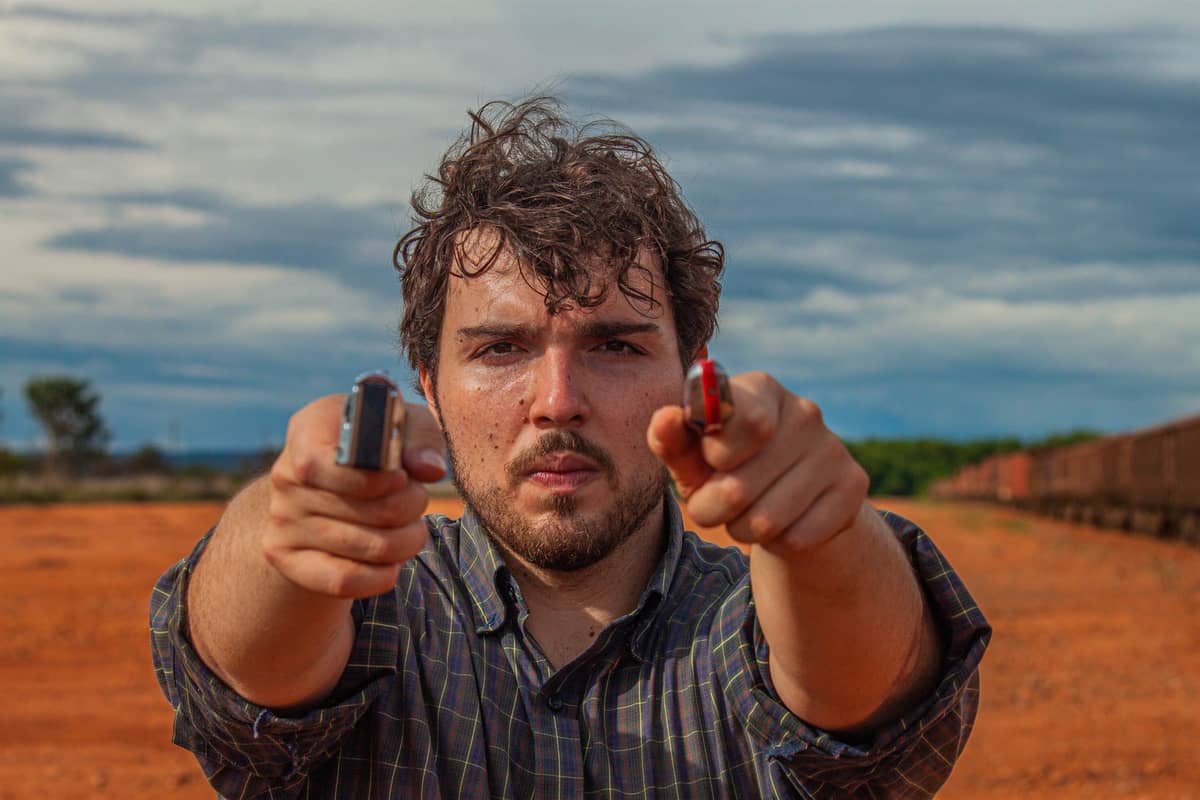 Guilherme Barros apontando duas gaitas diatônicas para a câmera como se fossem revólveres, na terra vermelha ao lado de um trem de carga